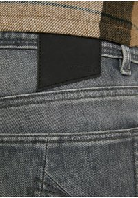 Grå denimjeans med en svart läderlapp märkt "CHASIN,'" med framträdande sömmar och en texturerad finish. Lager med ett mönstrat tyg.