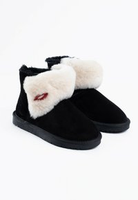 Botines de ante negros con un puño de piel sintética en tono crema, que presentan una suela texturizada y un pequeño detalle bordado en rojo en la piel.