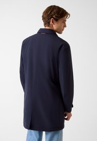 Marineblauwe lange jas van soepel materiaal, met een klassieke kraag, rugventilatie en knoopmanchetten voor een verstelbare pasvorm.