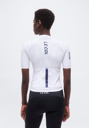 Inimene, kes kannab valget jalgrattasärki, millel on taga ja varrukal tekst "LE COL", ühendatuna mustade jalgrattapükstega, millel on "LE COL" vöökoht.