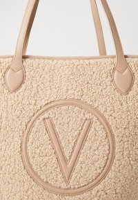 Beige tote-väska med en texturerad, ludden yta och släta läderdetaljer. Har en framträdande cirkulär logotyp med ett "V" i mitten.
