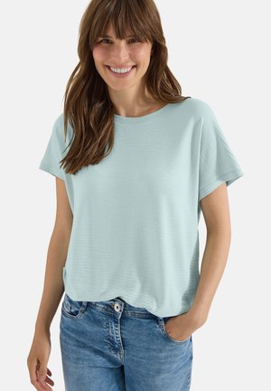 Femme souriante aux longs cheveux bruns, portant un haut texturé bleu clair à manches courtes et un jean bleu, une main dans la poche.