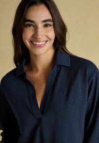 Naeratav naine, kellel on pikad tumedad juuksed, kannab tumesinist kaelusega särki, mille taustal on hallikasbeez taust.