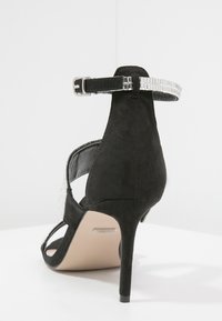 Sandale à talon haut en velours noir avec un talon fin, une sangle de cheville ornée de métal argenté et un design ouvert à l'arrière. Accentuée par un détail en strass.