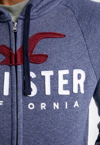 Zip hoodie tillverkad av mjuk, strukturerad marinblått tyg. Har vit och röd broderad text samt en känguruficka.