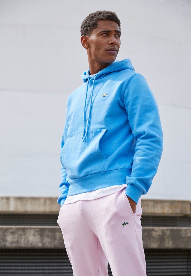 Sweat à capuche bleu avec une poche kangourou et des cordons, associé à un pantalon de survêtement rose clair portant un logo vert.