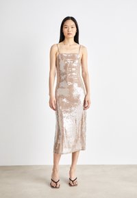 Αμάνικη, μεσαίου μήκους φόρεμα σε λαμπερή μπεζ παγιέτα, με τετράγωνη λαιμόκοψη και με λεπτή υφή; φοριέται με απλές μαύρες σάντες.