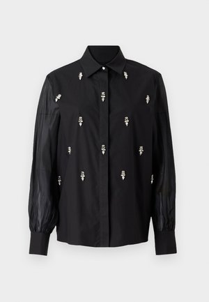 Sort skjorte med en struktureret design, der har gennemsigtige ærmer og sølvfarvede dekorative detaljer placeret lodret på forsiden.