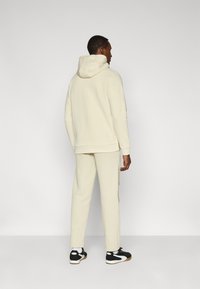 Μπεζ φούτερ με κουκούλα, σετ με τζόγκερς στο ίδιο μπεζ χρώμα. Διαθέτει ριμπ μανσέτες και άνετη εφαρμογή, συνδυασμένο με μαύρα και λευκά αθλητικά παπούτσια.