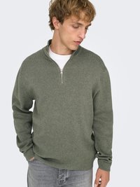 Pull vert clair à fermeture éclair avec un col montant, en tissu texturé. Porté sur un t-shirt blanc, avec des manches longues et une coupe décontractée.