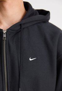 Sweatshirt preto com fecho éclair, feito de um tecido macio. Apresenta um logo branco da Nike na área superior esquerda do peito e um fecho de metal com uma aba para puxar.