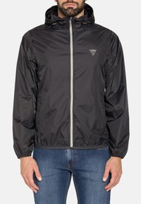 Giacca leggera nera con cappuccio, zip frontale, polsini elastici e patch con logo. Tessuto liscio e materiale resistente al vento.