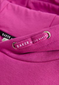 Felpa rosa con una texture liscia, cordino con un design intrecciato e un'etichetta del logo che legge "BAKER" vicino al collo.
