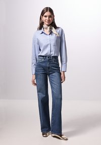 Blue striped button-up shirt with long sleeves, paired with high-waisted dark blue jeans and metallic gold flats. Model standing, hands at sides.