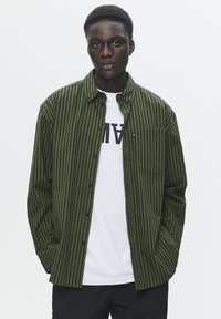 Groene en zwarte gestreepte button-up shirt met een borstzak, gelaagd over een wit T-shirt met grote letters, gedragen met zwarte broek.