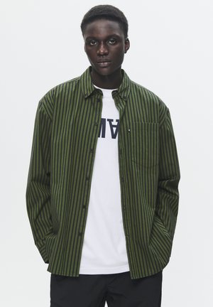 Green and black striped button-up shirt with a chest pocket, layered over a white T-shirt featuring bold lettering, worn with black pants.