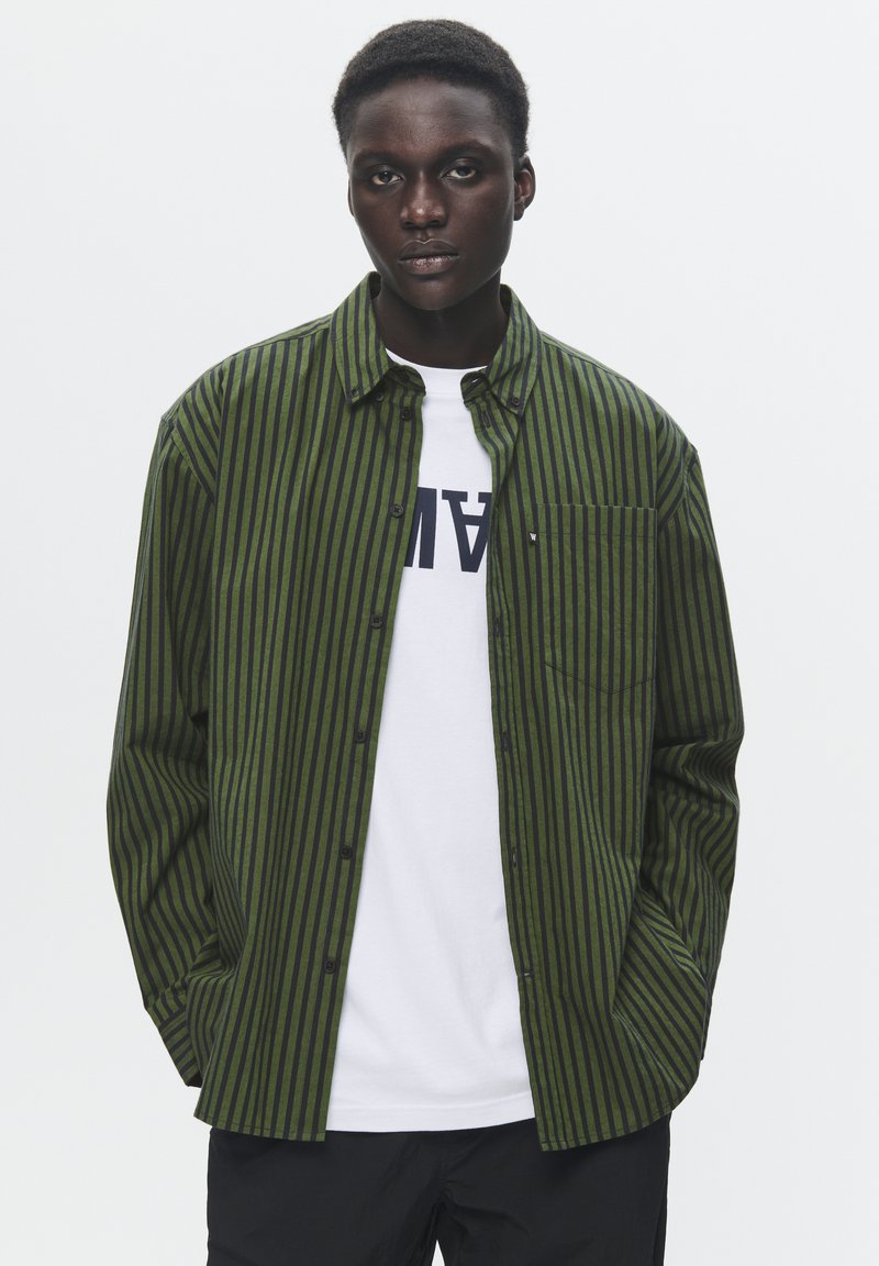 Groene en zwarte gestreepte button-up shirt met een borstzak, gelaagd over een wit T-shirt met grote letters, gedragen met zwarte broek.