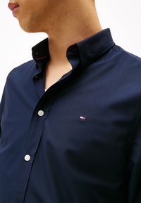 Camicia button-up blu navy con colletto strutturato e bottoni bianchi. Presenta un piccolo logo rosso, bianco e blu sul lato sinistro del petto.