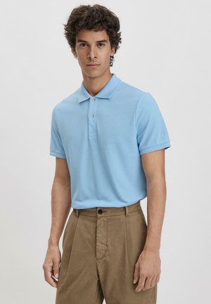 Himmelblaues Poloshirt aus strukturiertem Stoff, mit klassischem Kragen und zwei Knöpfen, kombiniert mit braunen Hosen.