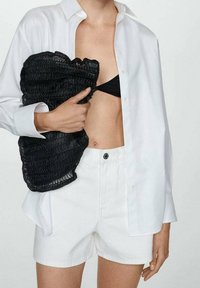 Camisa blanca de botones sobre un bikini negro, sosteniendo un chaleco negro texturizado; la modelo lleva puestos unos pantalones cortos blancos, mostrando una textura de piel suave.
