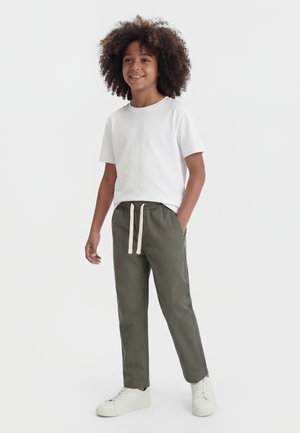 Dziecko z kręconymi włosami w białej koszulce, oliwkowych spodniach z troczkami i białych trampkach, stoi z jedną ręką w kieszeni.