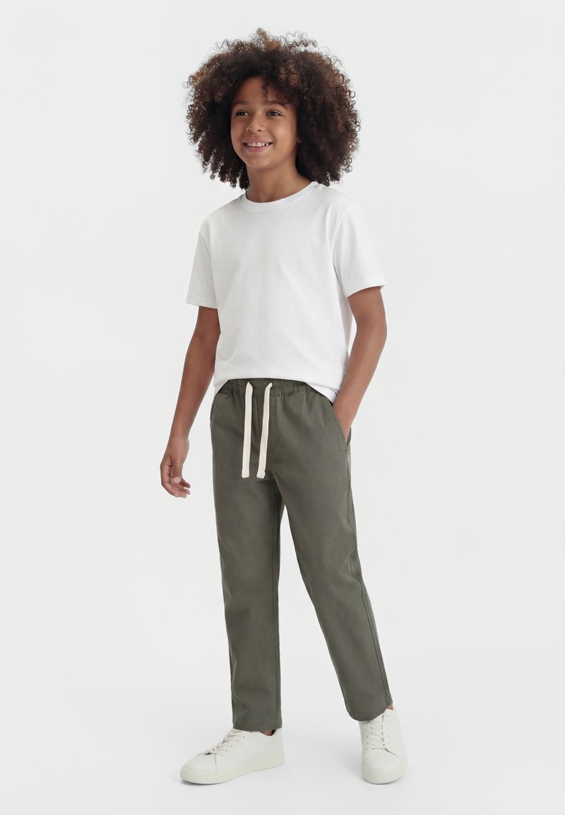 Vaikas su garbanotais plaukais, vilki baltą marškinėlį, alyvuogių žalių raištelių kelnes ir baltus sportinius batelius, stovintis su viena ranka kišenėje.