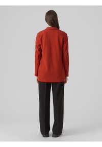 Långärmad, orange blazer med krage, med rak skärning. Matchas med svarta vida byxor. Texturerad tyg synlig.