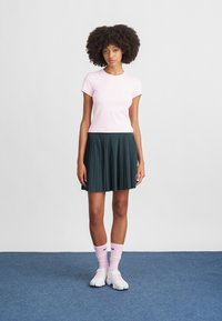 Jasnoróżowa bluzka z krótkim rękawem w połączeniu z plisowaną ciemnozieloną spódnicą. Modelka ma na sobie białe buty i różowe skarpetki Nike, stoi na niebieskim dywanie.