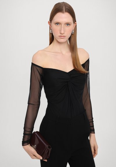 Melna bodysuit ar atklātiem pleciem, caurspīdīgām garām piedurknēm un sasietu priekšpusi. Modelis tur spīdīgu maroon krāsas somiņu. Vienkāršs fons.