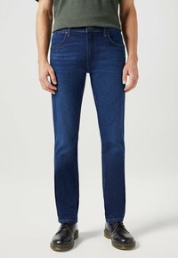 Vaqueros de mezclilla azul oscuro con corte recto, con bolsillos y cierre de botón estándar. Llevados con zapatos negros de cordones.