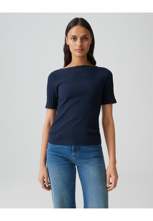 Femme portant un haut côtelé bleu marine à manches courtes et un jean bleu, debout devant un fond gris clair uni, faisant face à l'objectif.