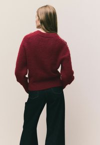 Röd fluffig tröja med långa ärmar och nedsänkta axlar, prydd med dekorativa detaljer på axeln, tillsammans med mörka jeans.