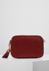 Kompaktowa prostokątna, ciemnoczerwona skórzana torebka z złotym łańcuchem i detalem w postaci frędzla, umieszczona na białej powierzchni na jednolitym tle.