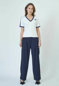 Camiseta blanca de cuello en V con ribete azul marino y logotipo, combinada con pantalones sueltos azul marino. Tejido suave con un corte relajado y pliegues sutiles.