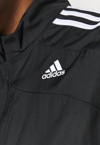 Giacca nera in tessuto testurizzato con colletto alto, tre strisce bianche sulla spalla e logo Adidas bianco.