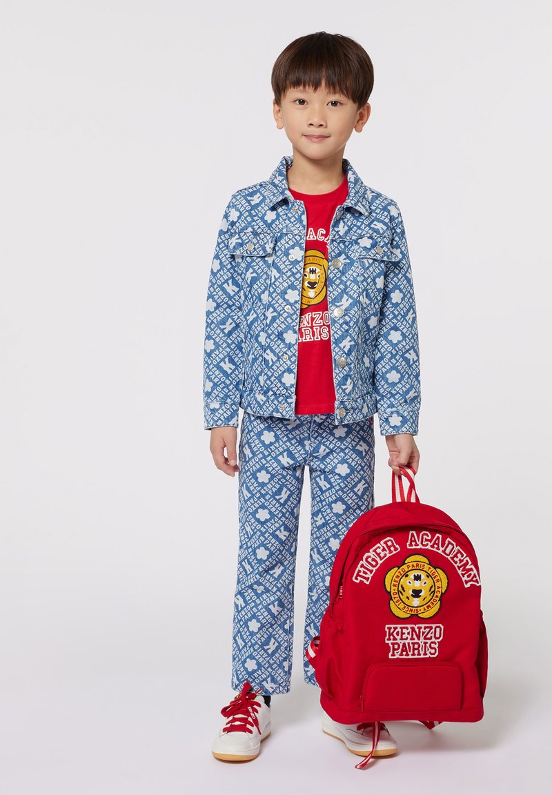 Giacca di denim blu e pantaloni con motivo bianco su tutta la superficie, t-shirt rossa con logo, zaino rosso con dettagli ricamati, sneakers bianche.