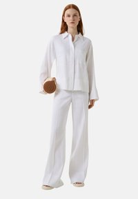 Witte linnen blouse met lange mouwen en een zak, gecombineerd met witte wijde broek. Model houdt een ronde tan crossbody tas vast.