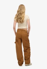 Pantalones cargo marrones hechos de una tela duradera, con múltiples bolsillos, corte relajado y un diseño de cintura alta. Parte superior beige simple visible.