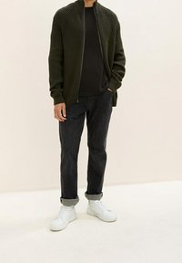Olivgrön ribbad zip-up cardigan med hög krage, bärs över en svart skjorta. Matchas med mörka jeans och vita sneakers.