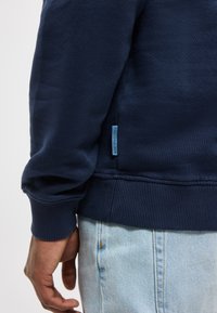 Αριστερό χέρι και μπράτσο άνδρα με μπλε σκουρόχρωμο φούτερ δίπλα σε ανοιχτό μπλε τζιν με ορατό πλάγιο ραφή και μικρή ετικέτα υφάσματος.