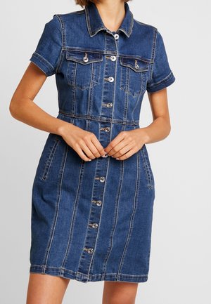 Kvinde iført en kortærmet denimkjole med knapper foran, brystlommer og lodrette sømdetaljer.