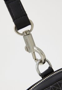 Fekete bőrszíj ezüstszínű fém csattal, amelyen "Calvin Klein" felirat látható, egy fekete cipzáros táskához csatlakoztatva, amelynek texturált díszítése van.