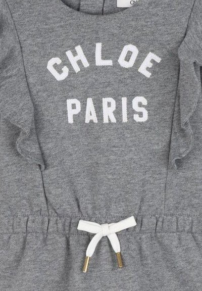 Szara dzianinowa odzież z białym napisem "CHLOE PARIS", z falbaniastymi detalami po bokach oraz białym sznurkiem związanym w kokardkę w talii.
