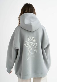 Felpa oversize grigia chiara con un disegno a linee bianche di un volto e fiori sulla parte posteriore. Tessuto morbido con cappuccio e polsini a coste.