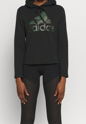 Schwarzer, kurzer Adidas-Hoodie mit grünem Camouflage-Logo, getragen mit schwarzen Leggings mit Netzmuster an einer stehenden Person.