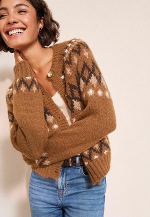 Kvinna som ler och har på sig en brun mönstrad stickad kofta över en vit topp och blå jeans med örhängen, stående mot en beige vägg.