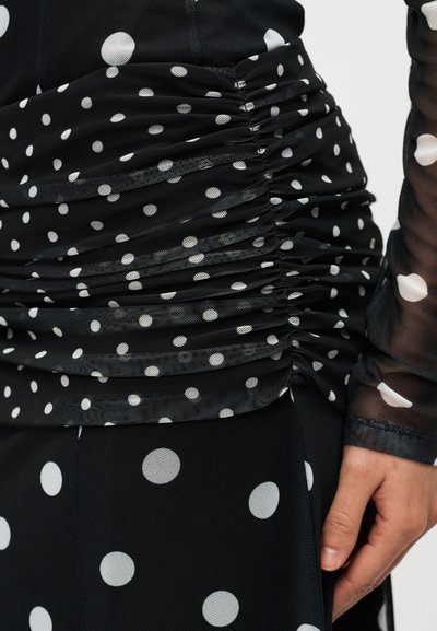 Μαύρο τοπ από ύφασμα με υφή ρυμουλκούμενου, διακοσμημένο με λευκές πουά. Η σχεδίαση περιλαμβάνει διαφανή μανίκια και εφαρμοστή σιλουέτα.