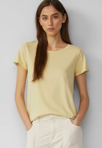 Lichtgeel T-shirt met korte mouwen van zachte stof met een ronde halslijn. Het model poseert met de handen in de zakken, en draagt crèmekleurige broek.