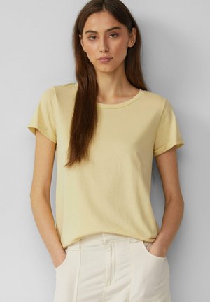 T-shirt jaune clair à manches courtes en tissu doux avec un col rond. Le mannequin pose les mains dans les poches, portant un pantalon couleur crème.