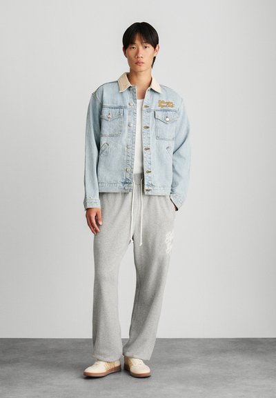 Lichtblauwe denimjas met beige kraag, zakken en gouden borduurwerk, gecombineerd met grijze sweatpants en beige sportschoenen.